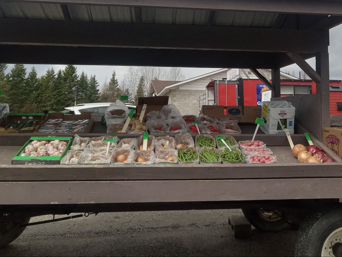 A rustic outdoor market stall on a trailer with a red roof, displaying fresh produce including green beans, red tomatoes, onions, and garlic. A cardboard box labeled 'HARDY 80L' is visible on the right side, with trees and a cloudy sky in the background.