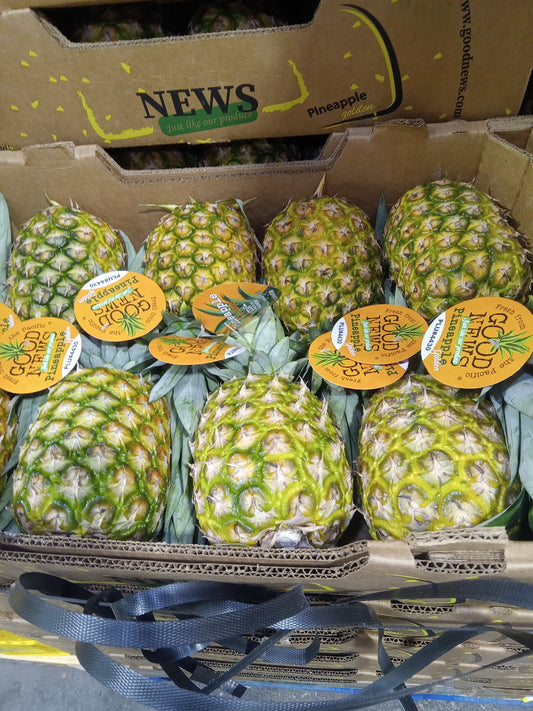 Several whole pineapples with attached yellow circular stickers are displayed in a cardboard shipping crate. A larger box behind them shows the brand name NEWS Pineapple.